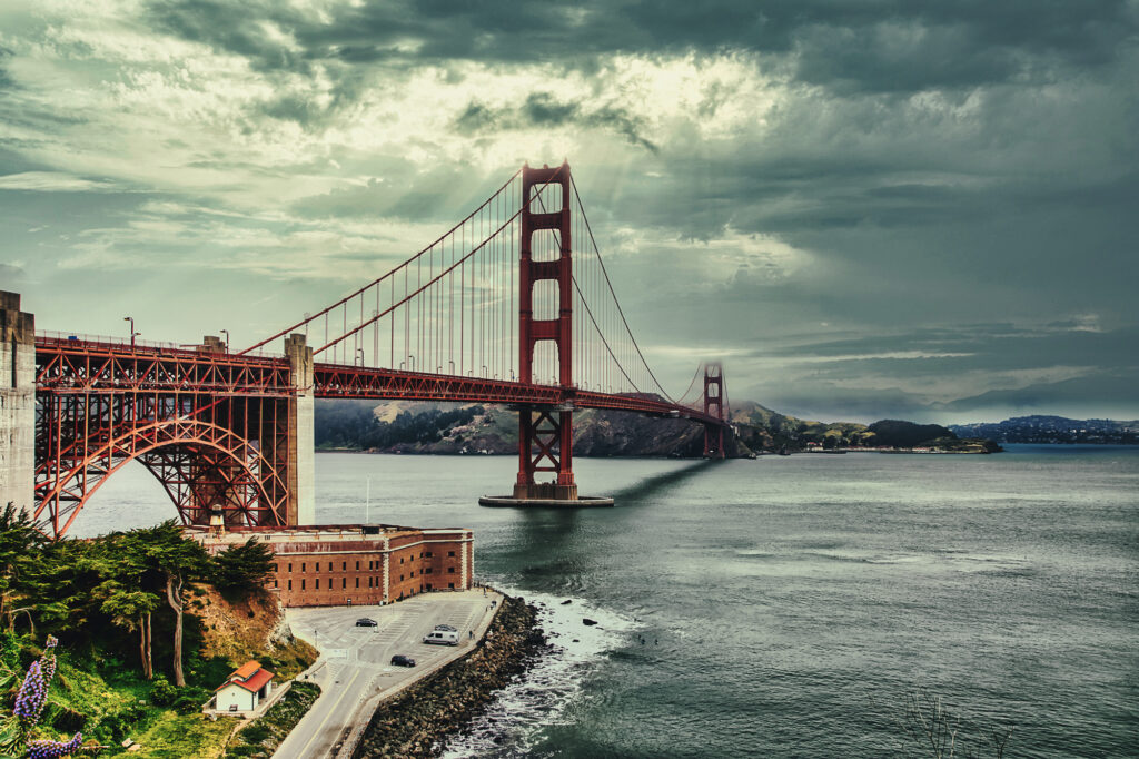 A photo of the Golden Gate Bridge under cloudy skies. Sunrays are popping through the clouds. On the left of the frame is the structures that the road sits on. Across the cam waters of the bay the Marin Headlands can be seen.